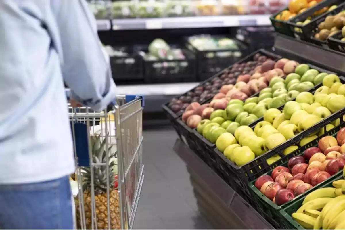 Persona empujando un carrito de compras en un supermercado frente a una sección de frutas con manzanas y plátanos.