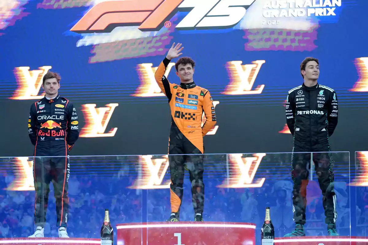 Three racing drivers on the podium of the 2023 Australian Grand Prix, with the driver in the center raising his hand in greeting. Three racing drivers on the podium of the 2023 Australian Grand Prix, with the driver in the center raising his hand in greeting.