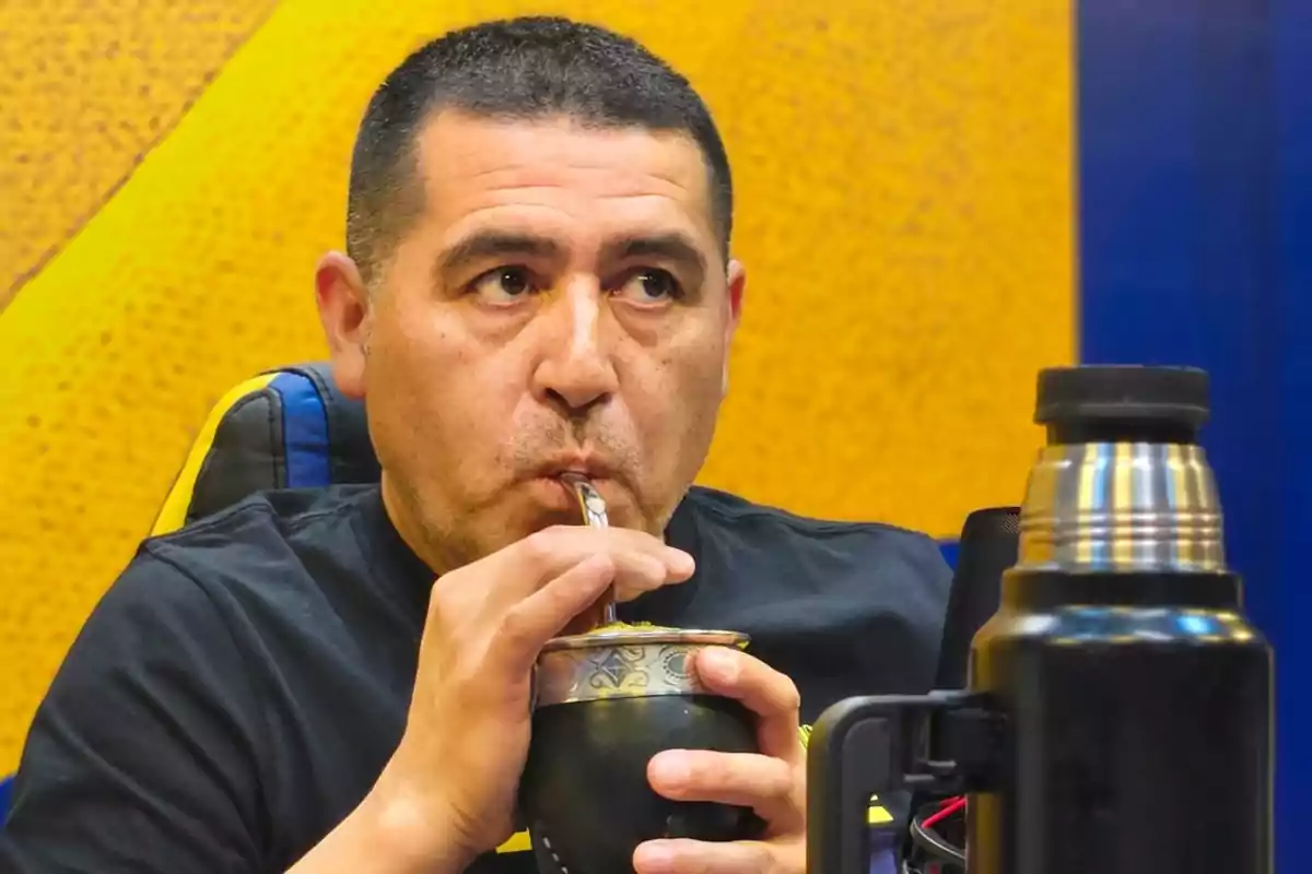 Hombre de cabello corto tomando mate con una bombilla, sentado frente a un fondo amarillo y azul, con un termo negro en primer plano.