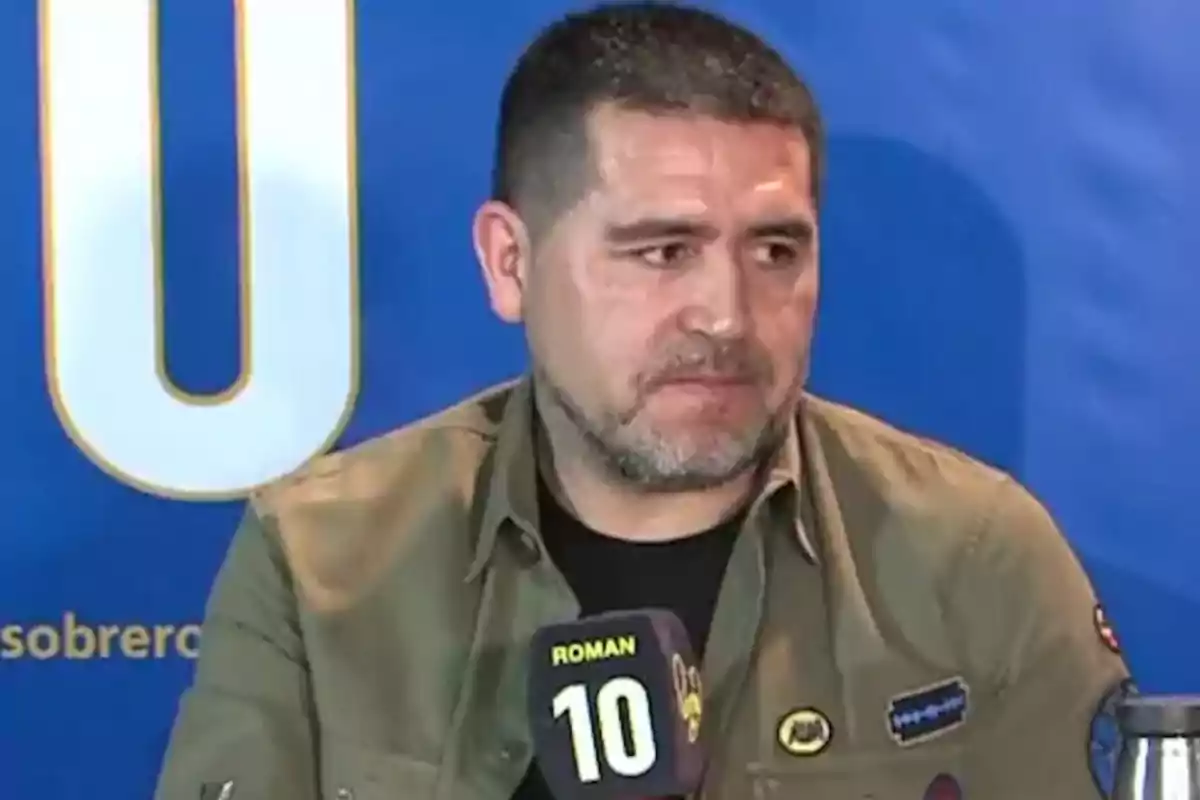 A man with a beard and short hair is sitting in front of a microphone with the number 10 on a blue background. A man with a beard and short hair is sitting in front of a microphone with the number 10 on a blue background.