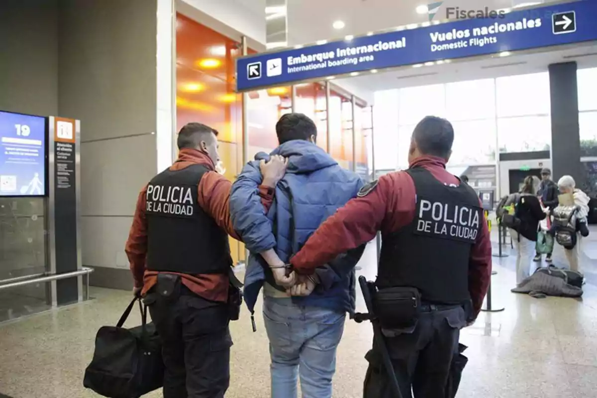 En la imagen se observa a dos policías de la ciudad escoltando a una persona esposada en un aeropuerto, dirigiéndose hacia la zona de embarque internacional.