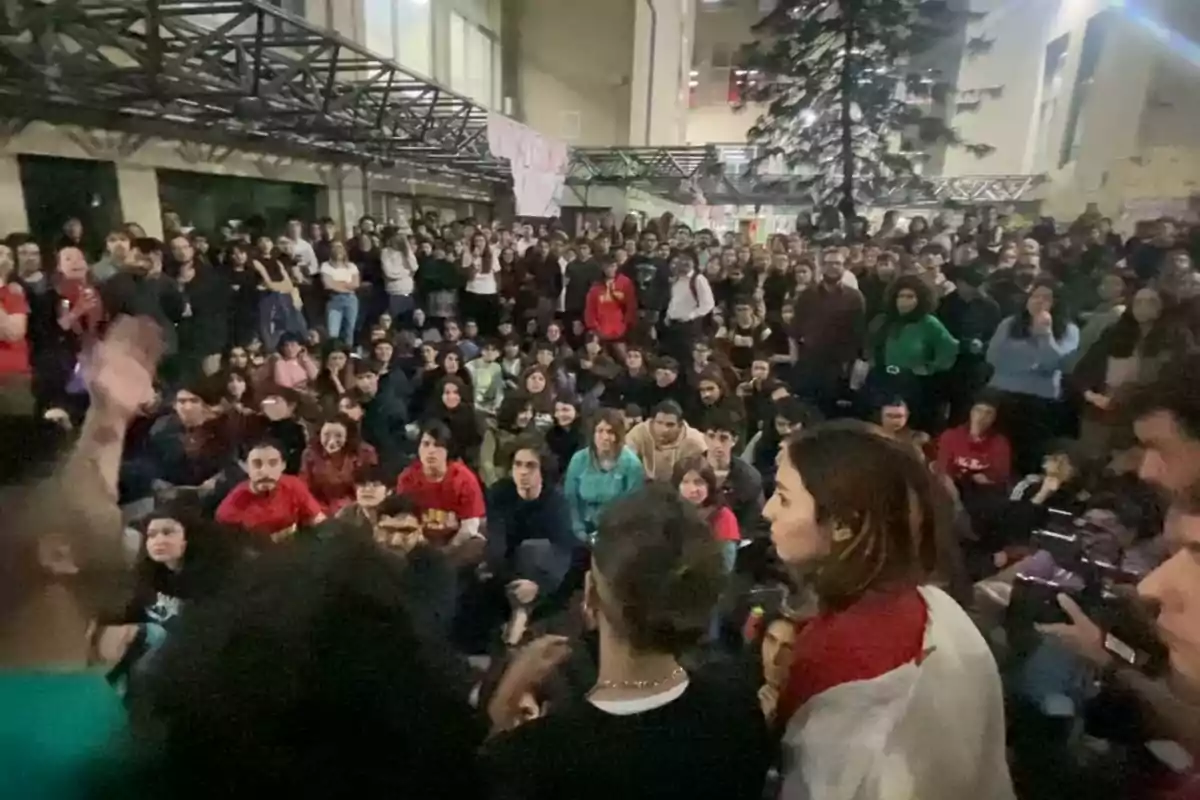 Un grupo numeroso de personas se encuentra reunido en un espacio interior, algunas están sentadas mientras otras permanecen de pie, observando atentamente hacia el frente.