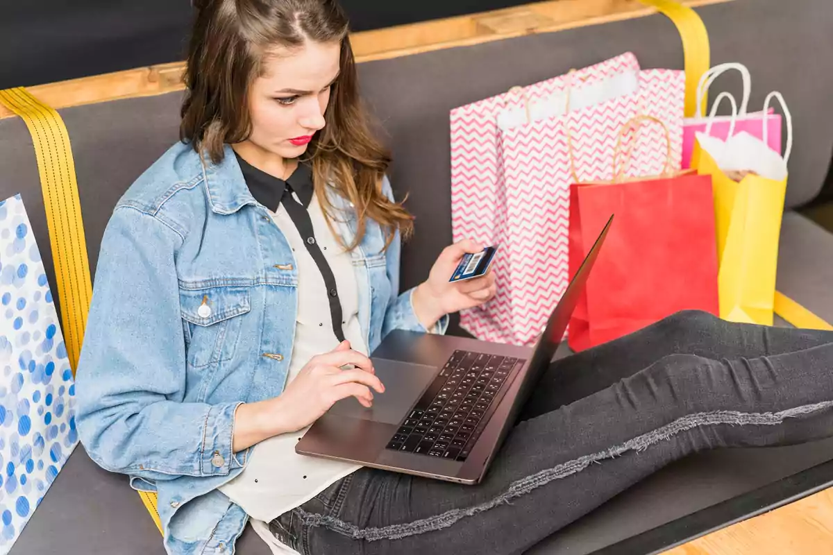 Mujer sentada en un sofá usando una laptop con bolsas de compras a su alrededor.