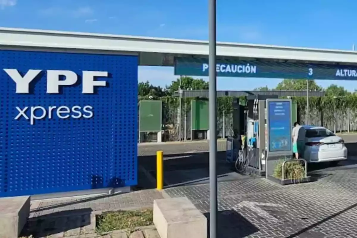 Estación de servicio YPF xpress con un auto blanco cargando combustible en un surtidor y un cartel azul grande a la izquierda Estación de servicio YPF xpress con un auto blanco cargando combustible en un surtidor y un cartel azul grande a la izquierda
