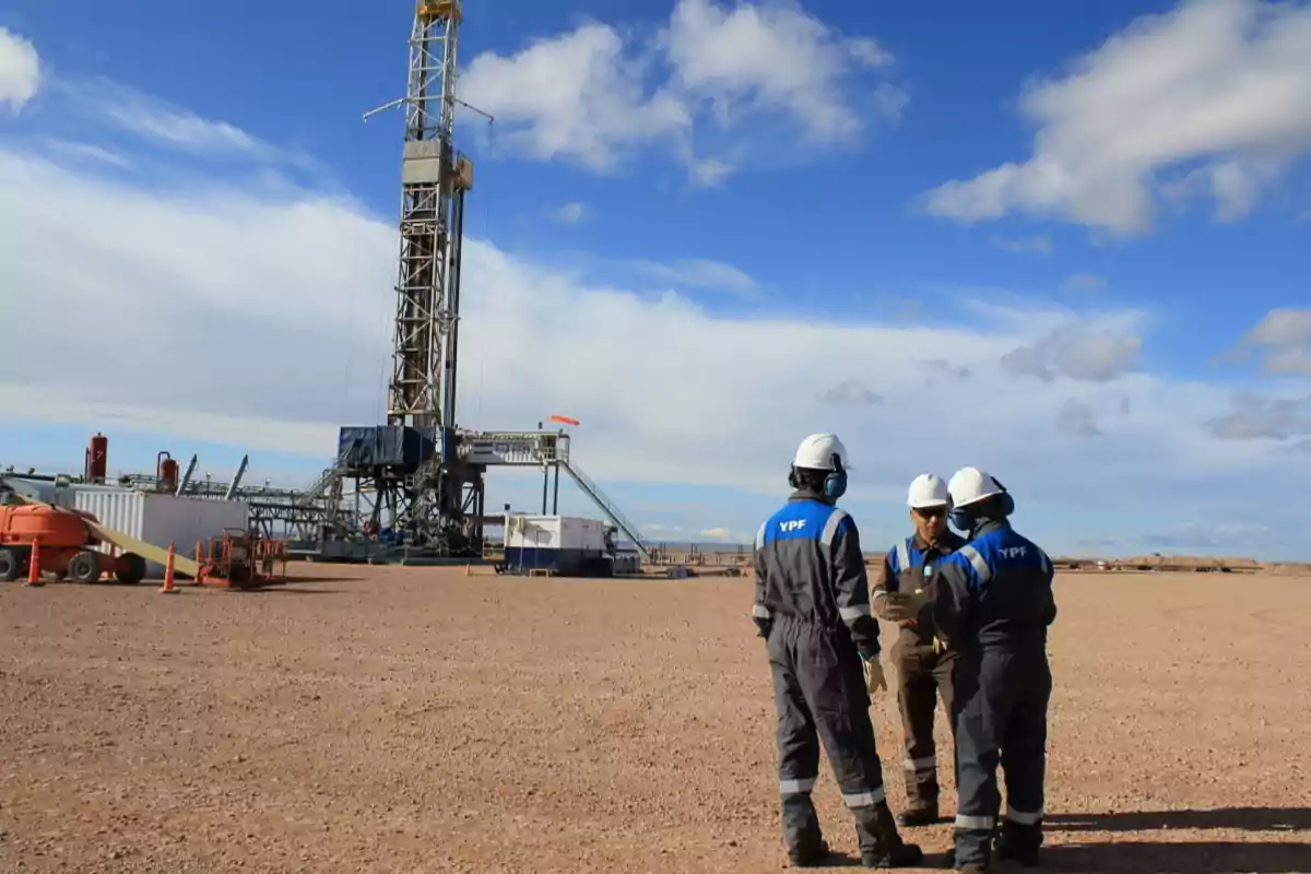 Trabajadores con cascos y uniformes de YPF conversan cerca de una torre de perforación en un campo petrolero bajo un cielo azul.