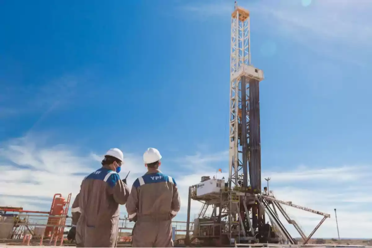 Dos trabajadores con cascos observan una plataforma de perforación petrolera bajo un cielo despejado.