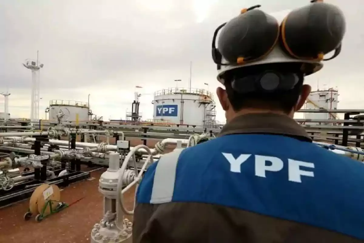 YPF worker with his back turned in uniform at an industrial plant with tanks and pipes.