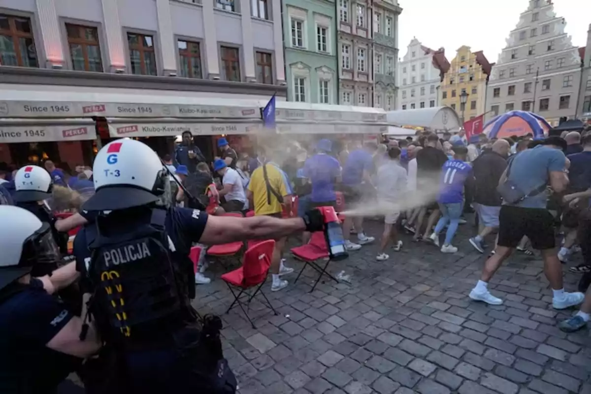 Un policía con casco y chaleco rocía gas pimienta hacia un grupo de personas en una plaza con edificios antiguos y terrazas llenas de gente