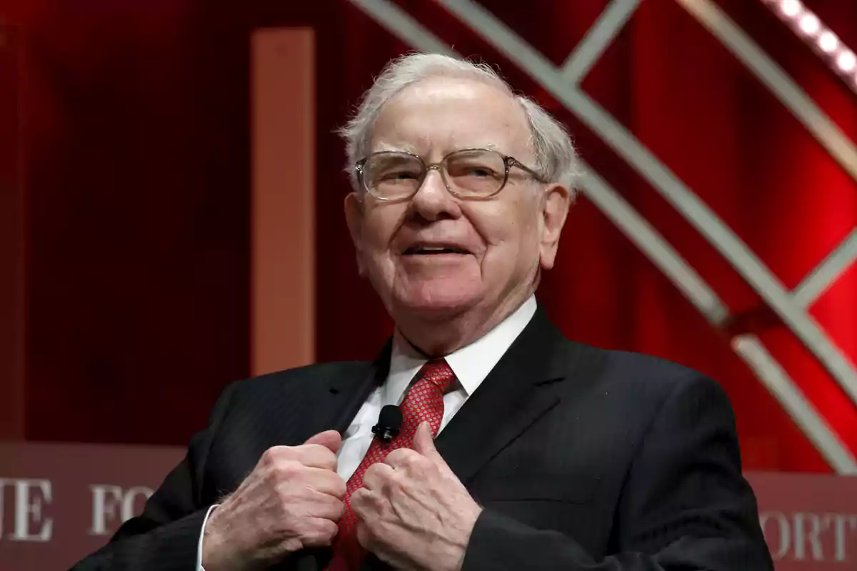 An older man with glasses and a dark suit smiles while adjusting his red tie on a stage with a red background.
