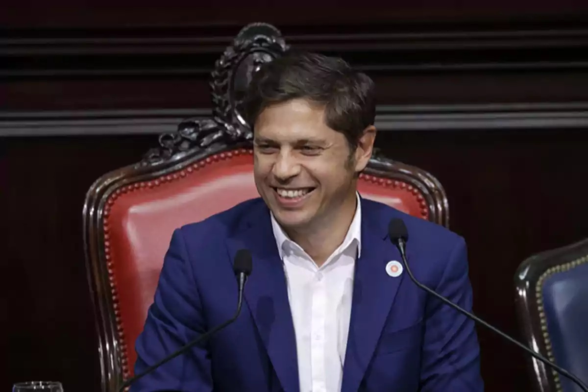 Un hombre sonriente con traje azul sentado en una silla con respaldo decorativo y dos micrófonos frente a él.