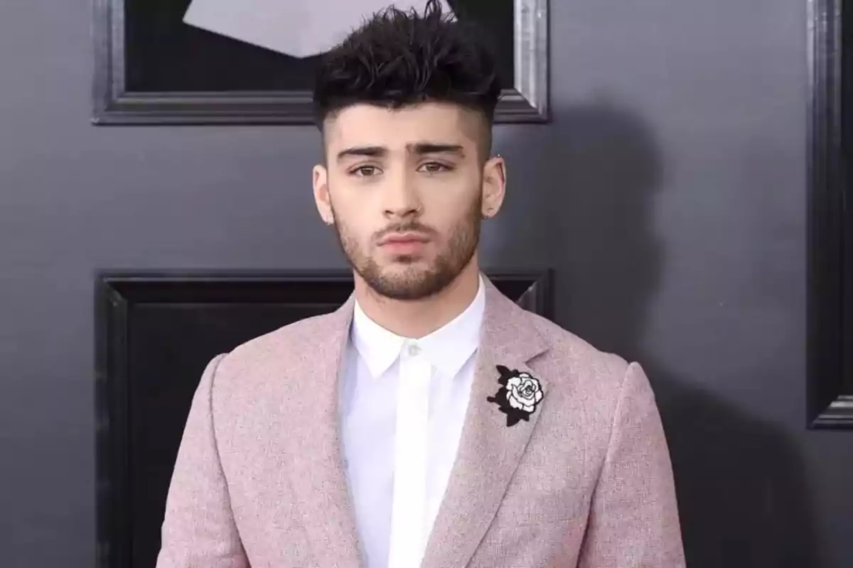 Un hombre con barba y cabello oscuro lleva un traje rosa claro con una camisa blanca y un broche de flor en la solapa.