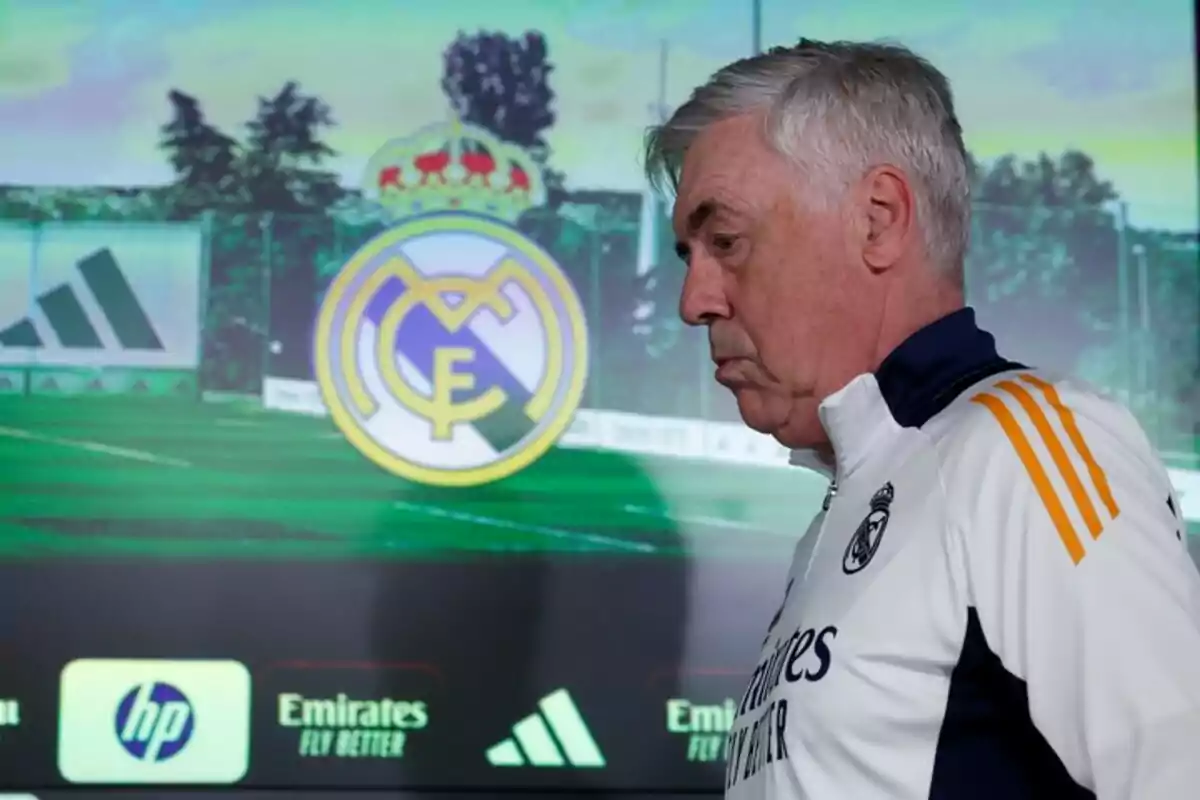 Un hombre con uniforme deportivo del Real Madrid está de perfil frente a un fondo con el escudo del club. Un hombre con uniforme deportivo del Real Madrid está de perfil frente a un fondo con el escudo del club.