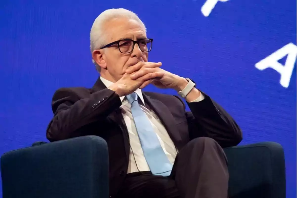 An older man with glasses and gray hair is sitting in an armchair with his hands clasped in front of his face, wearing a dark suit and a blue tie, against a blue background. An older man with glasses and gray hair is sitting in an armchair with his hands clasped in front of his face, wearing a dark suit and a blue tie, against a blue background.