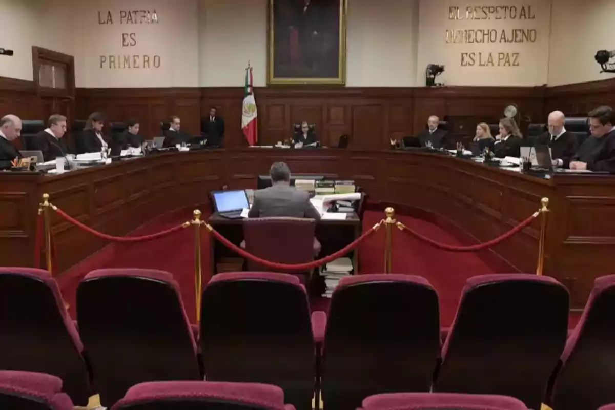 A courtroom with judges seated on a semicircular platform, a man in the center facing a table with documents and computers, and phrases on the wall that say A courtroom with judges seated on a semicircular platform, a man in the center facing a table with documents and computers, and phrases on the wall that say