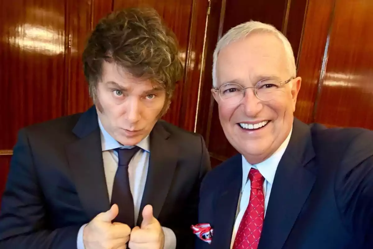 Two men dressed in suits pose for a selfie; one of them smiles while the other gives a thumbs-up gesture, both standing in front of a polished wooden wall. Two men dressed in suits pose for a selfie; one of them smiles while the other gives a thumbs-up gesture, both standing in front of a polished wooden wall.