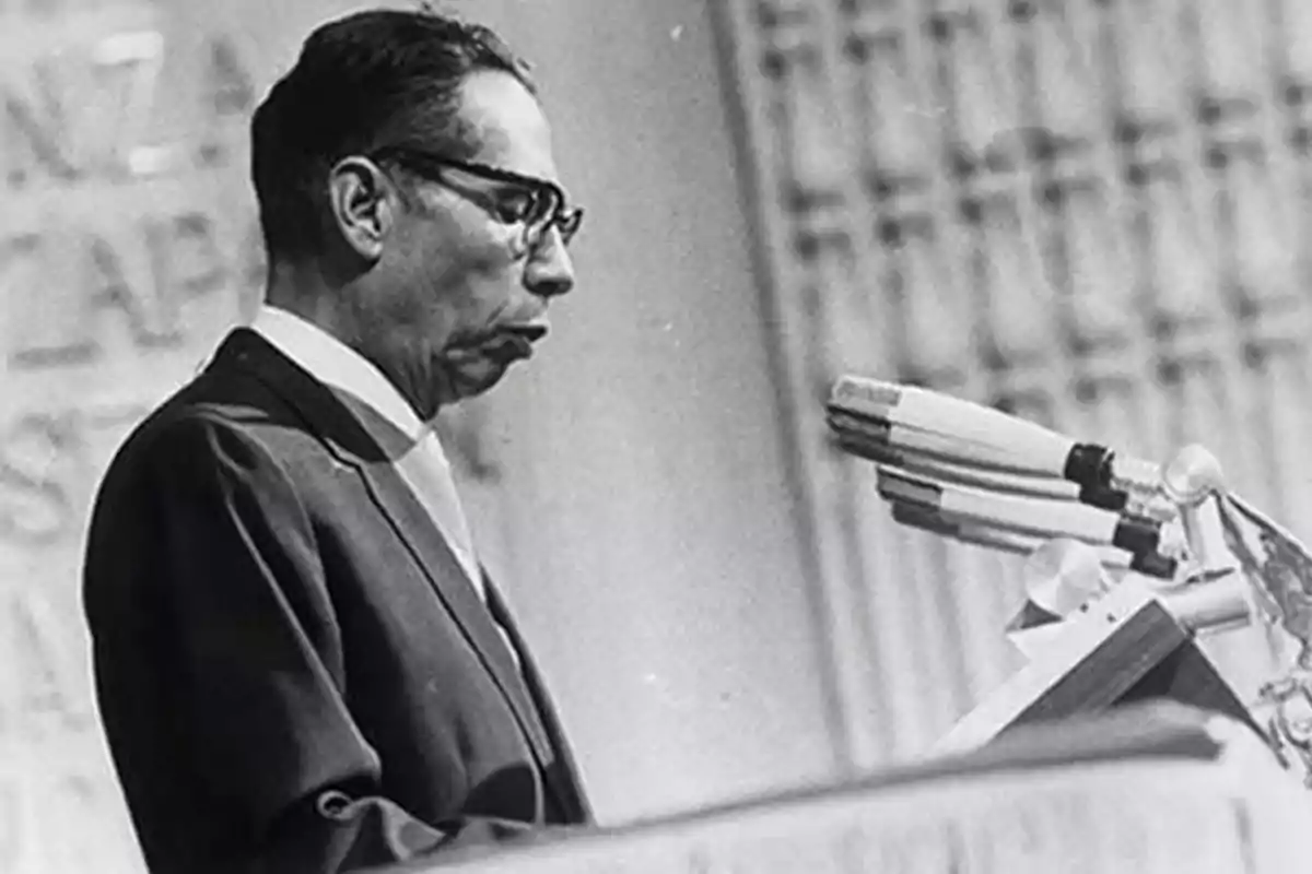 Man in a suit and glasses speaking in front of several microphones at a podium Man in a suit and glasses speaking in front of several microphones at a podium